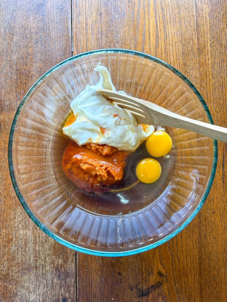 glass bowl on a kitchen table with a wooden spoon, pumpkin puree, eggs, and yogurt in it.