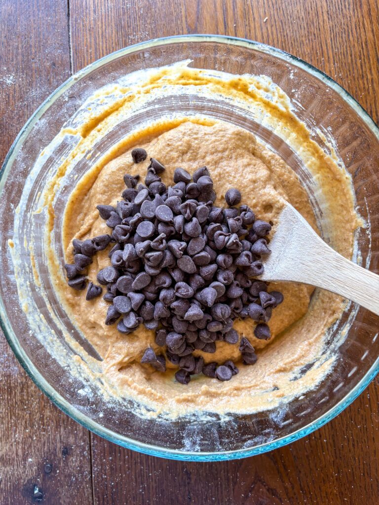 glass bowl with pumpkin muffin batter and chocolate chips and a wooden spoon on a kitchen table