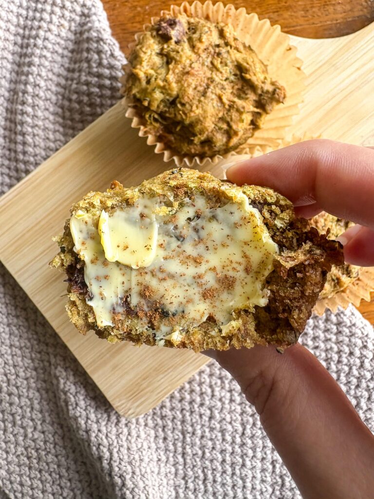 hand holding zucchini muffin with butter and cinnamon in front of a cutting board