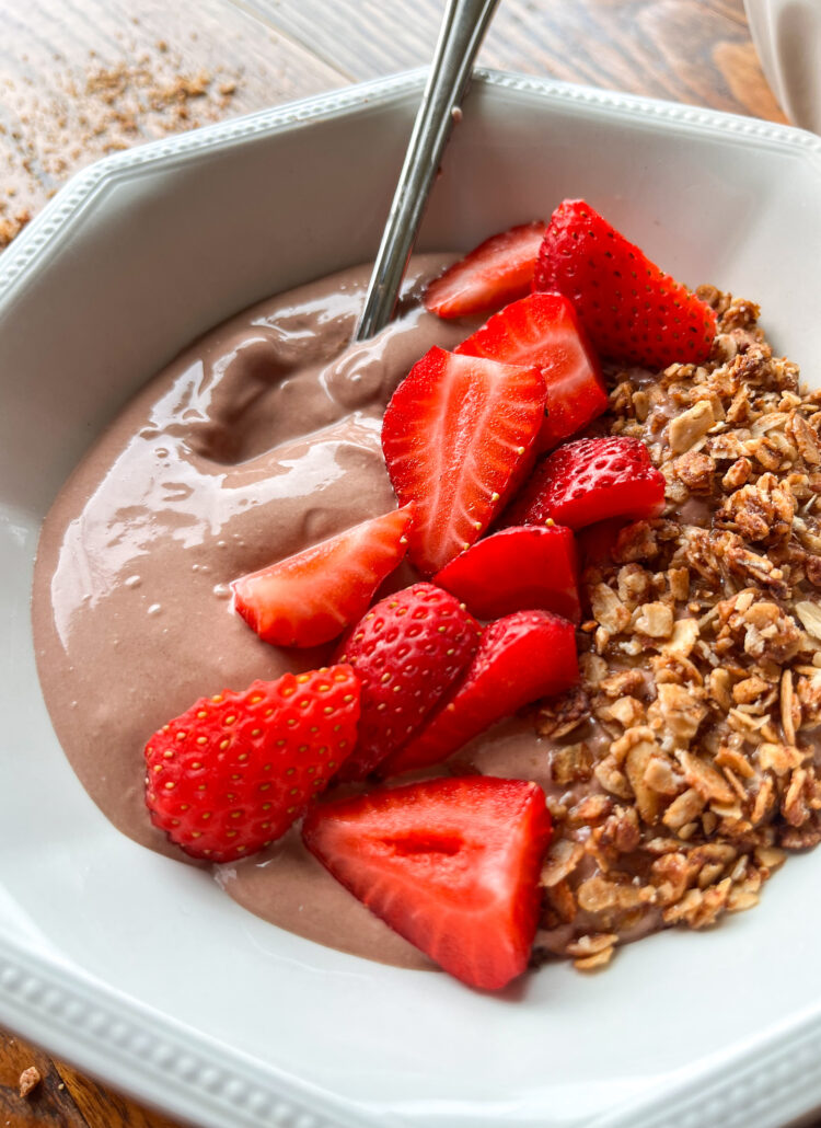 close up shot of chocolate whipped greek yogurt in a white bowl with a spoon, granola, and strawberries