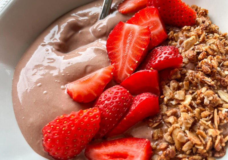 close up shot of chocolate whipped greek yogurt in a white bowl with a spoon, granola, and strawberries