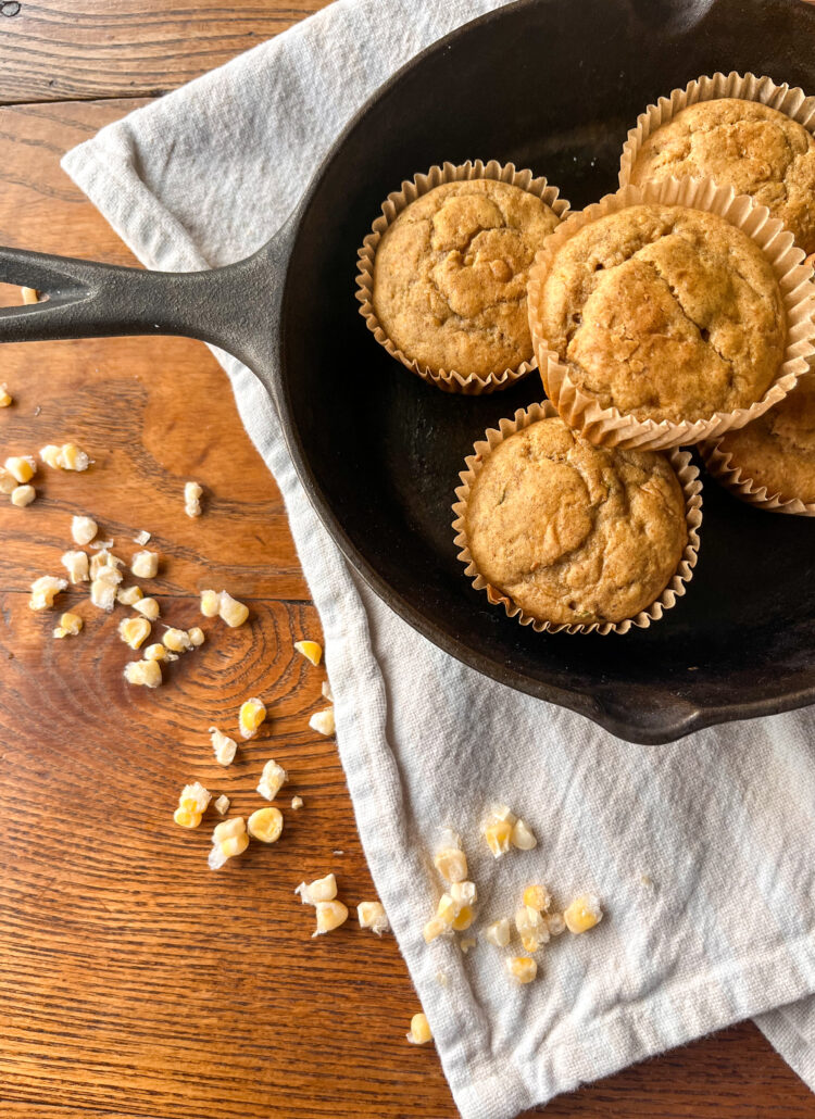 Cornbread muffins in a cast iron skillet with corn and a towel on a kitchen table