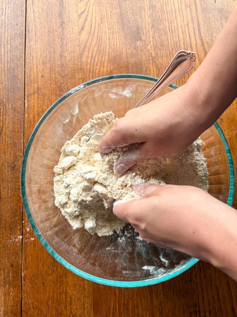 a woman's hand crumbling flour and butter together in a glass bowl on a kitchen table with a fork