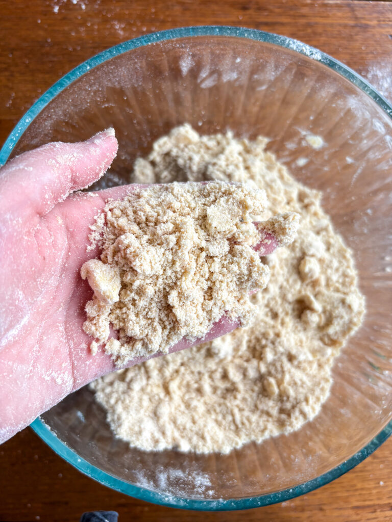 a hand holding flour and butter crumbles with flour and butter in a glass bowl in the background