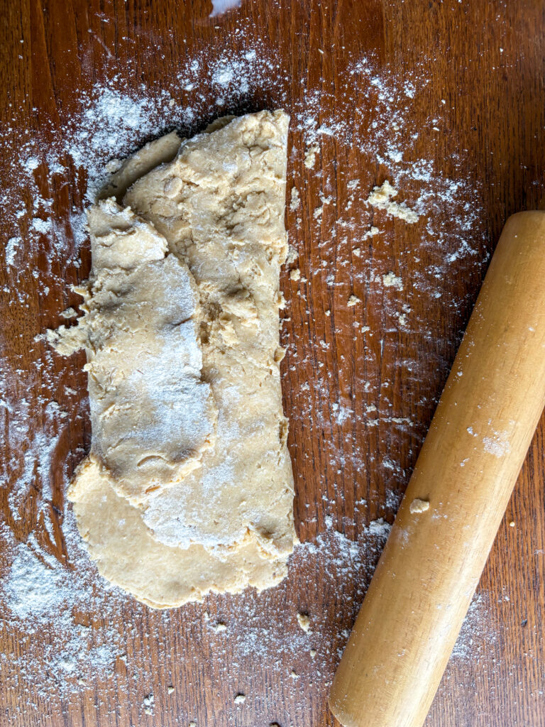 shaggy biscuit dough folded into a rectangle on a kitchen table with flour and a wooden rolling pin