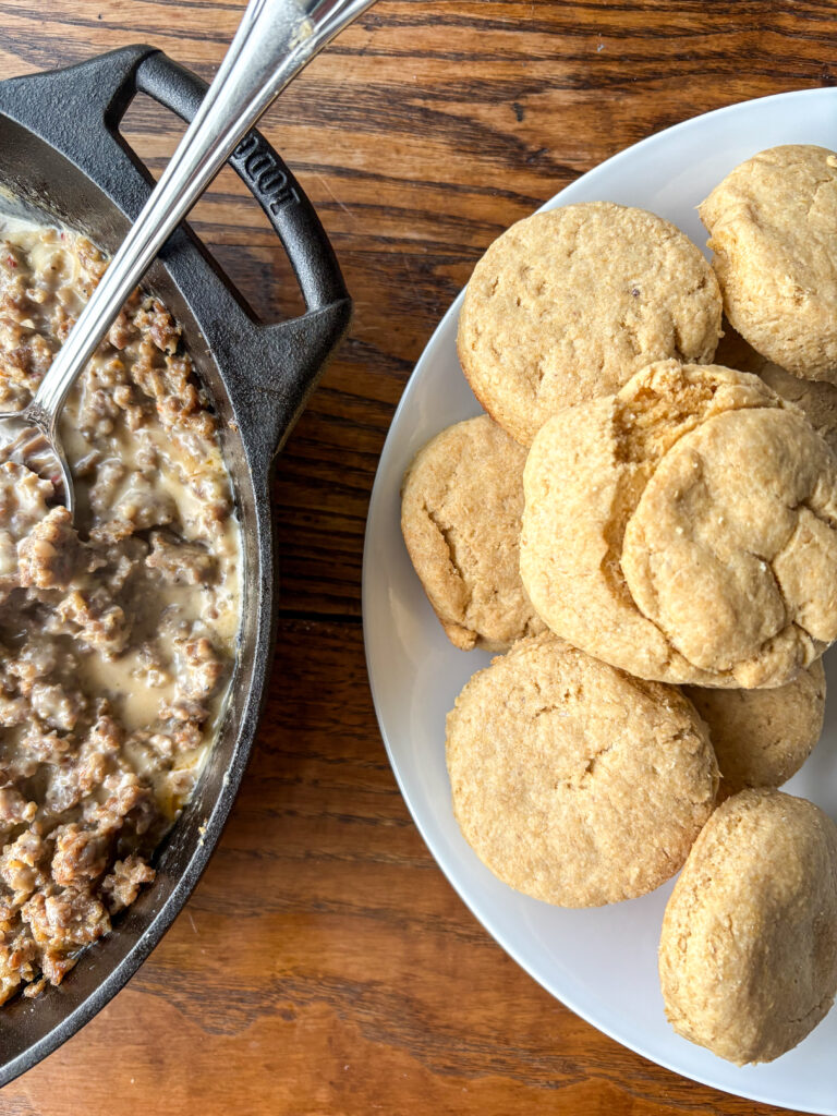 plate of biscuits on a kitchen table with cast iron skillet with sausage gravy and a spoon