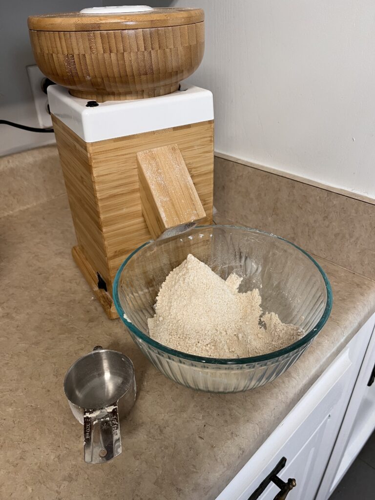 grain mill with a bowl of freshly milled flour on a countertop with a measuring cup. 