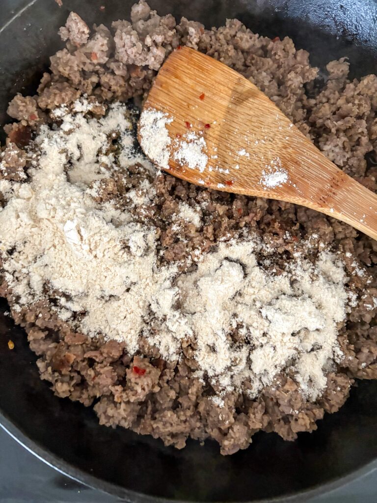 browned pork sausage in a cat iron skillet with a wooden spoon and fresh milled flour