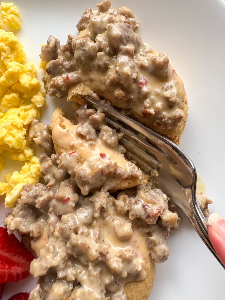 fresh milled flour biscuits and classic southern sausage gravy on a plate with eggs and a strawberry with a fork cutting into it