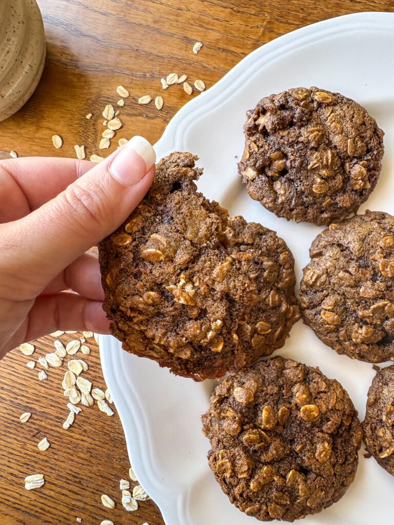 woman's hand holding a blackstrap molasses oatmeal cookie with a white plate and a kitchen table in teh background