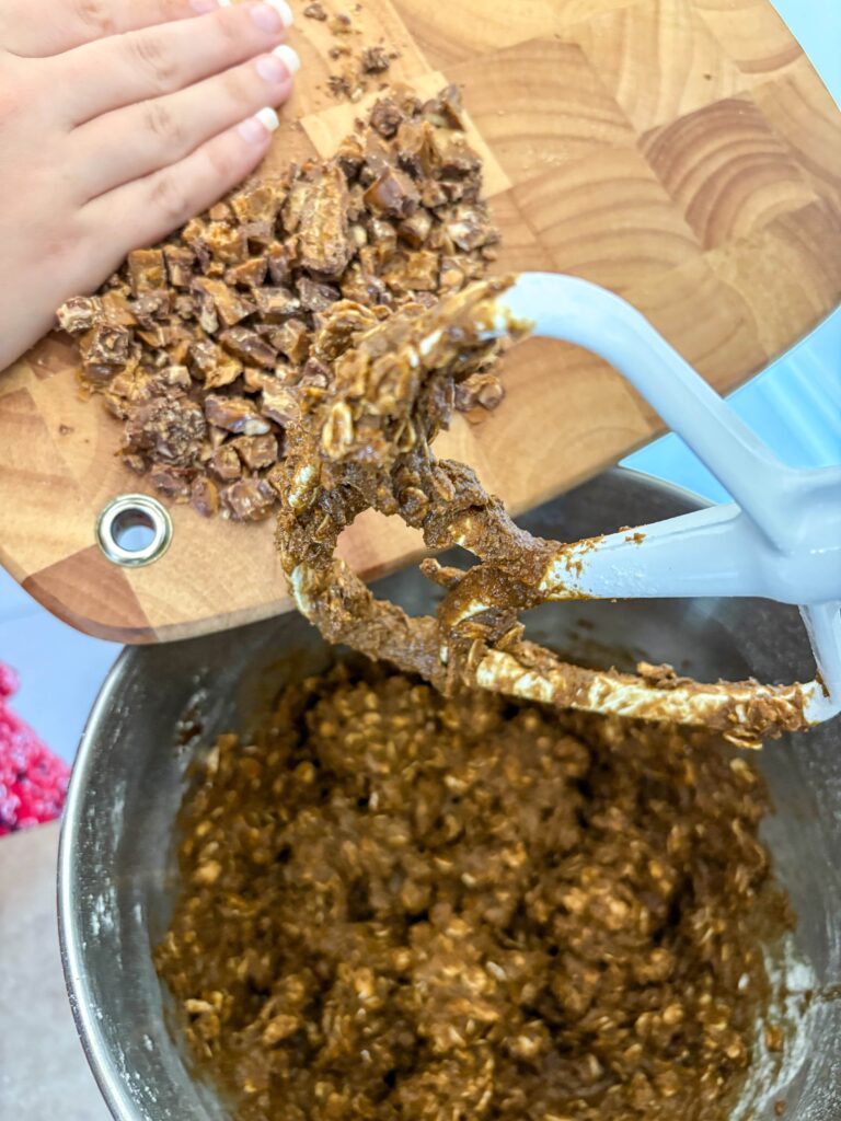 cutting board with a woman's hand pushing toffee bit into a bowl of blackstrap molasses oatmeal cookie dough with a stand mixer paddle attachment