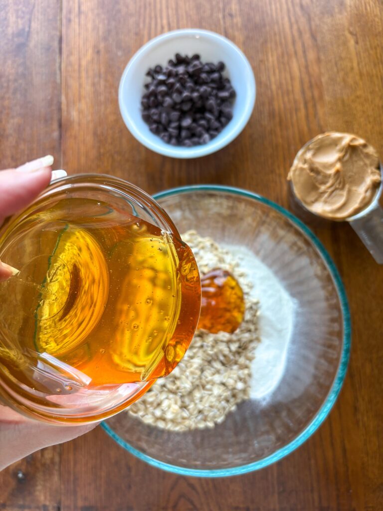 honey pouring into a bowl of oats on a kitchen table with peanut butter and chocolate chips