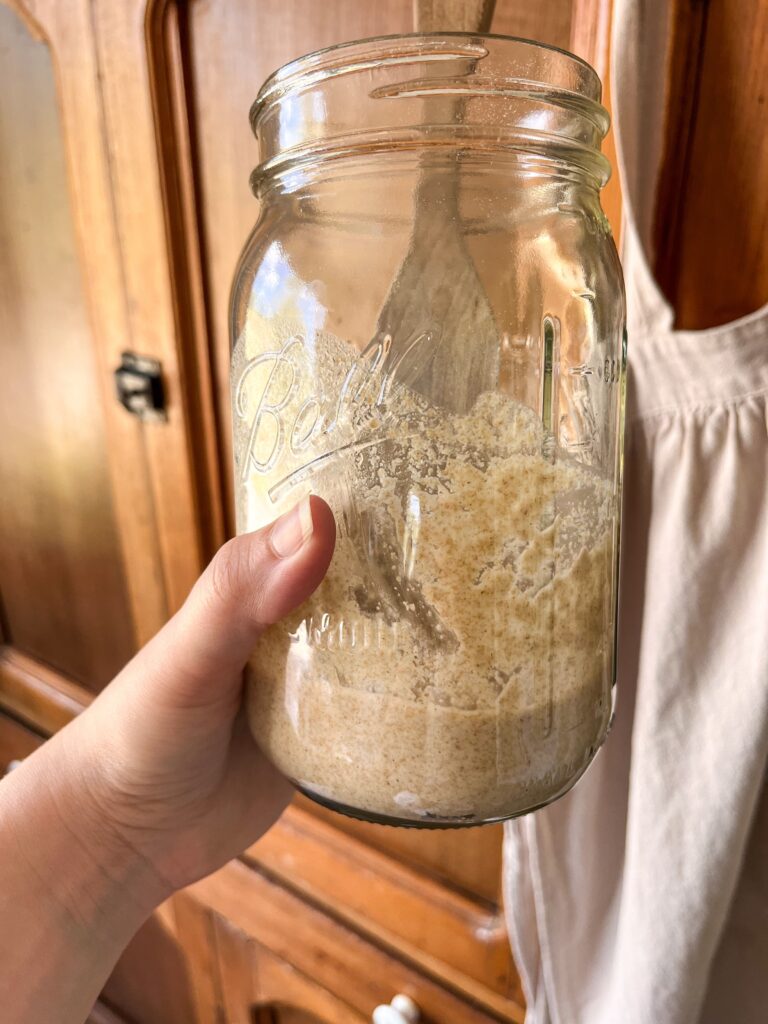 unfed sourdough starter in a mason jar with a wooden spurtle in front of a pie safe