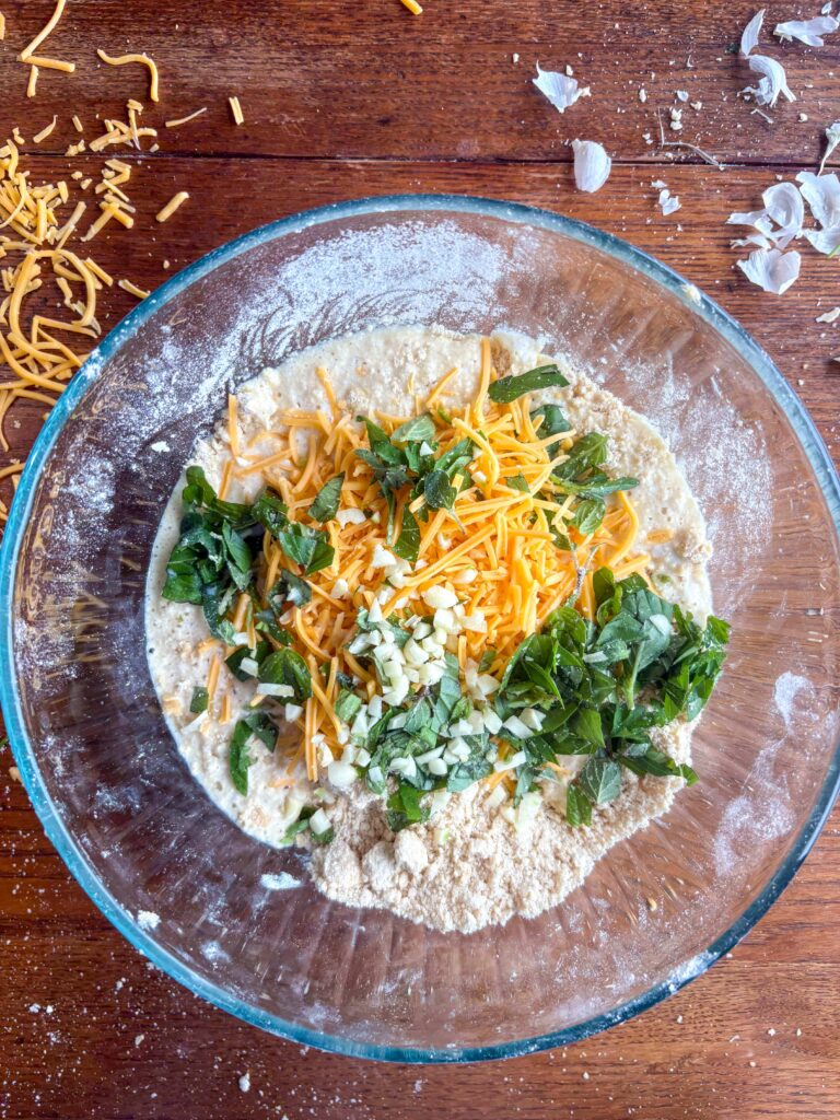 garlic cheddar biscuit dough in a glass bowl on a kitchen table with herbs, garlic, and cheese
